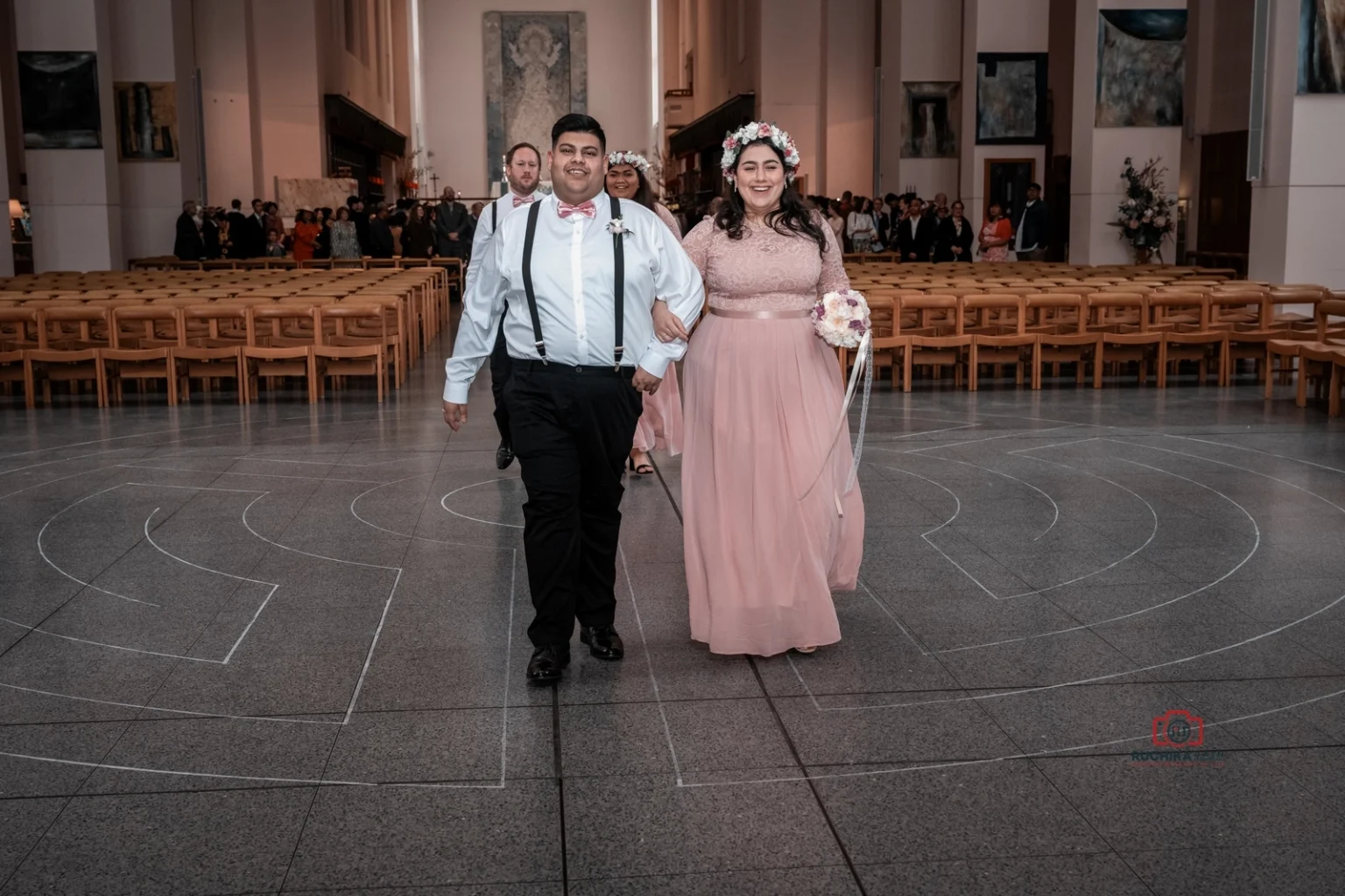 Bridesmaid in pink lace dress and groomsman in white shirt with suspenders walk arm-in-arm down the church aisle after the ceremony