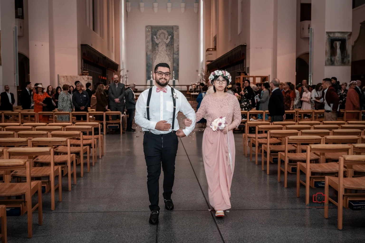 Bridesmaid in pink dress and floral crown walks arm-in-arm with groomsman in white shirt and suspenders down the church aisle