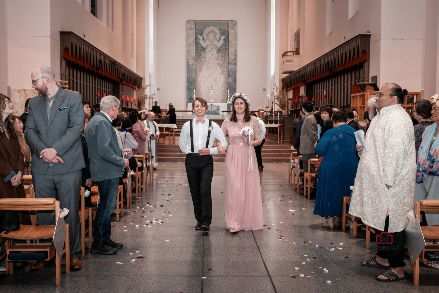 Bridesmaid in pink gown and floral crown walks arm-in-arm with groomsman wearing suspenders down the church aisle