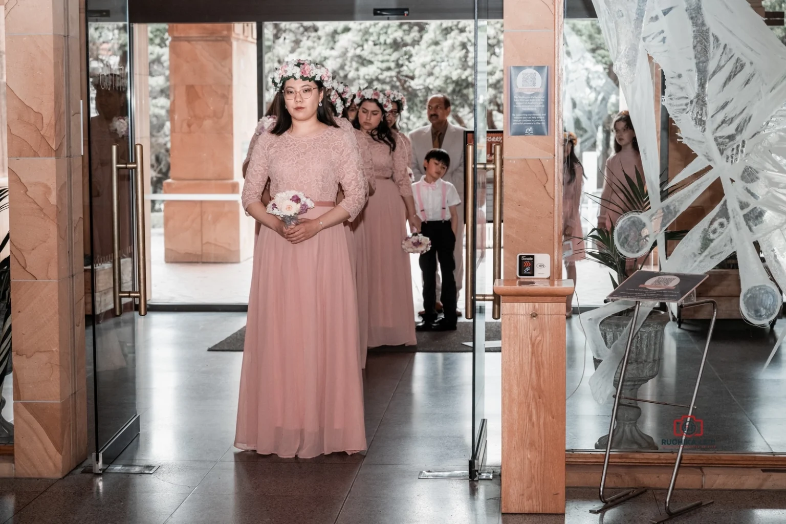 Bridesmaids in pink dresses and floral crowns enter the church, followed by a young ring bearer.