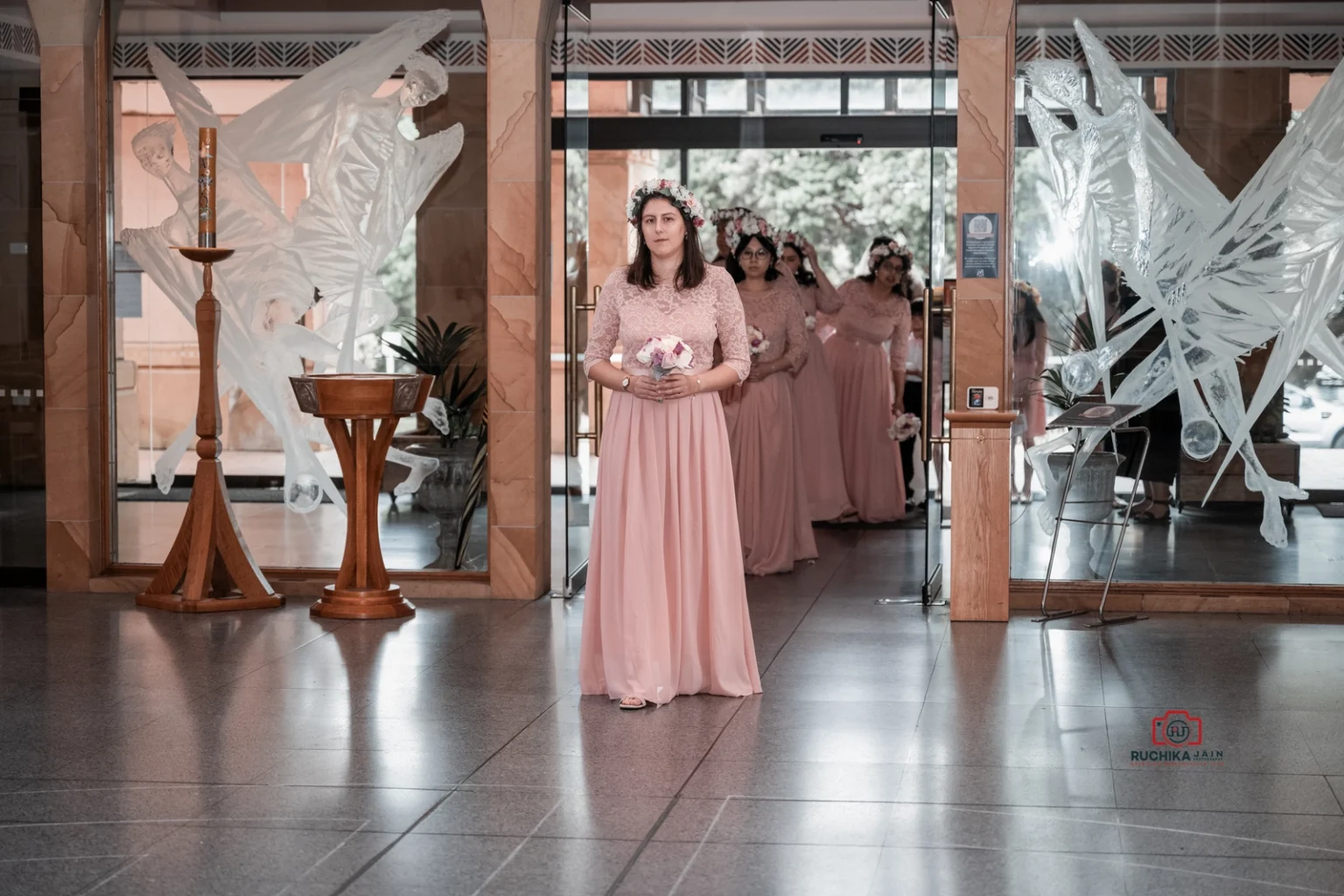 Bridesmaid in pink dress holding flowers leads others into a church, with glass decor in the background.