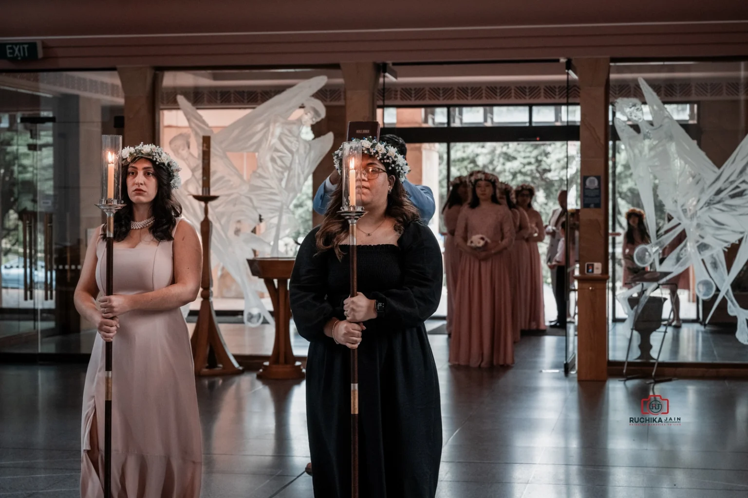 Two women in floral crowns holding candles lead a procession in a church, with bridesmaids in pink dresses following.