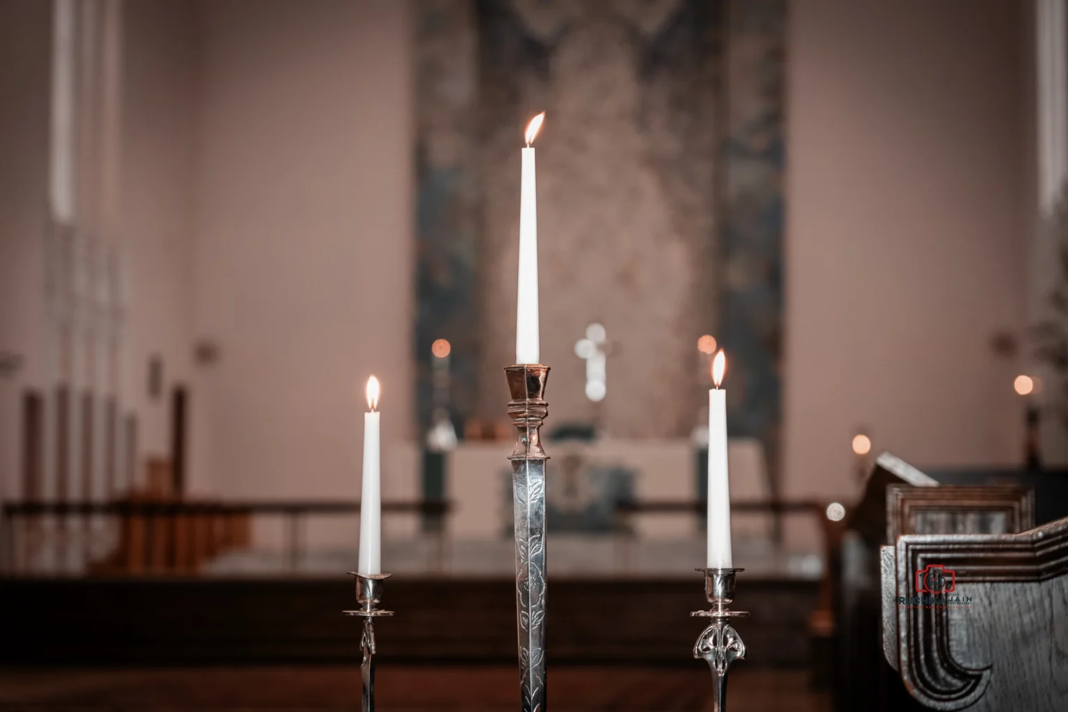 Three lit candles in silver holders on a church altar, with blurred pews and cross in the background.