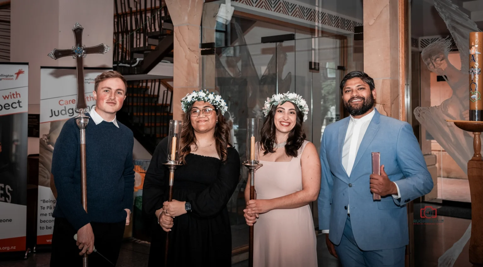 Four church ceremony attendants, two holding candles and one holding a cross, pose indoors, wearing floral crowns and formal attire.