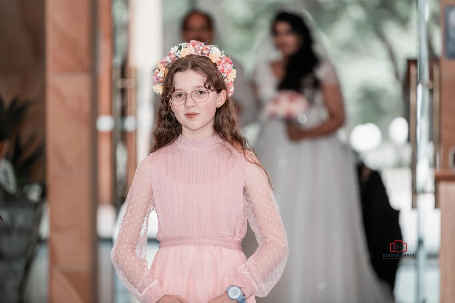 Young flower girl in a pink dress and floral crown walking down the aisle with the bride in the background.