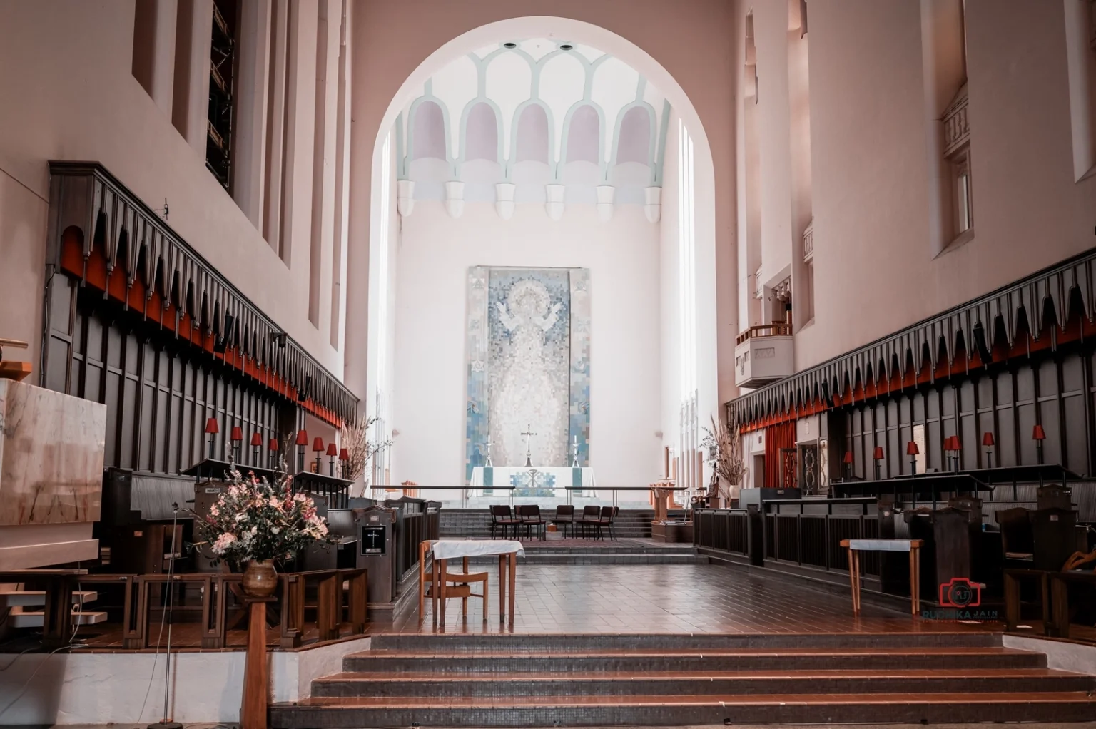 Interior view of a large church with high ceilings, arched altar area, and wooden pews, featuring a central artwork behind the altar