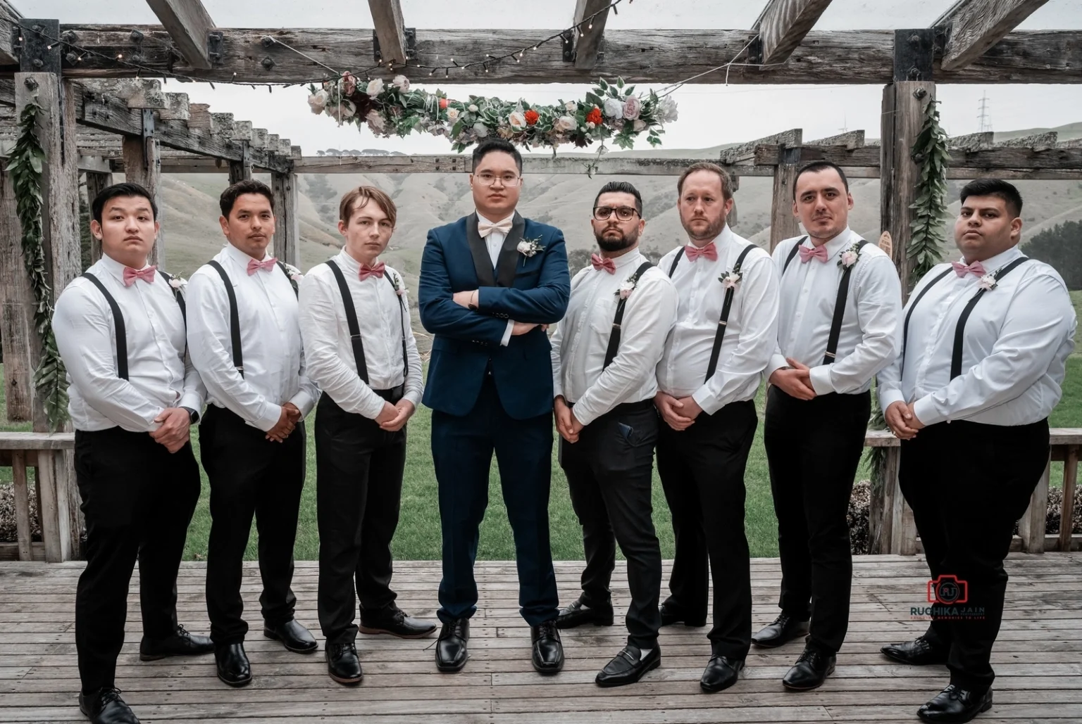 Groom in a blue suit stands with his groomsmen dressed in white shirts, black suspenders, and pink bow ties, posing seriously under a wooden pergola decorated with flowers