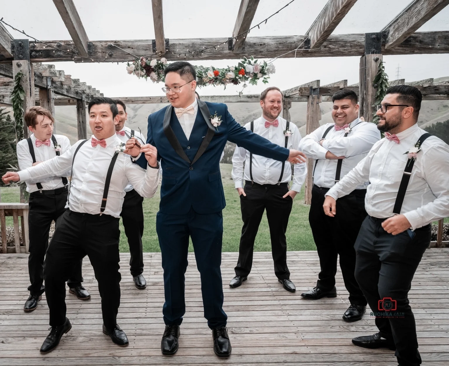 Groom and his groomsmen laughing and posing playfully under a wooden pergola decorated with flowers and greenery