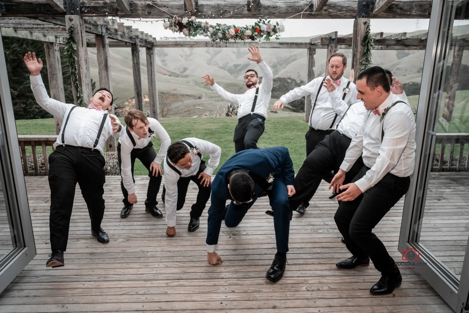 Groom and groomsmen posing in humorous action stances under a decorated wooden pergola, laughing and having fun outdoors.