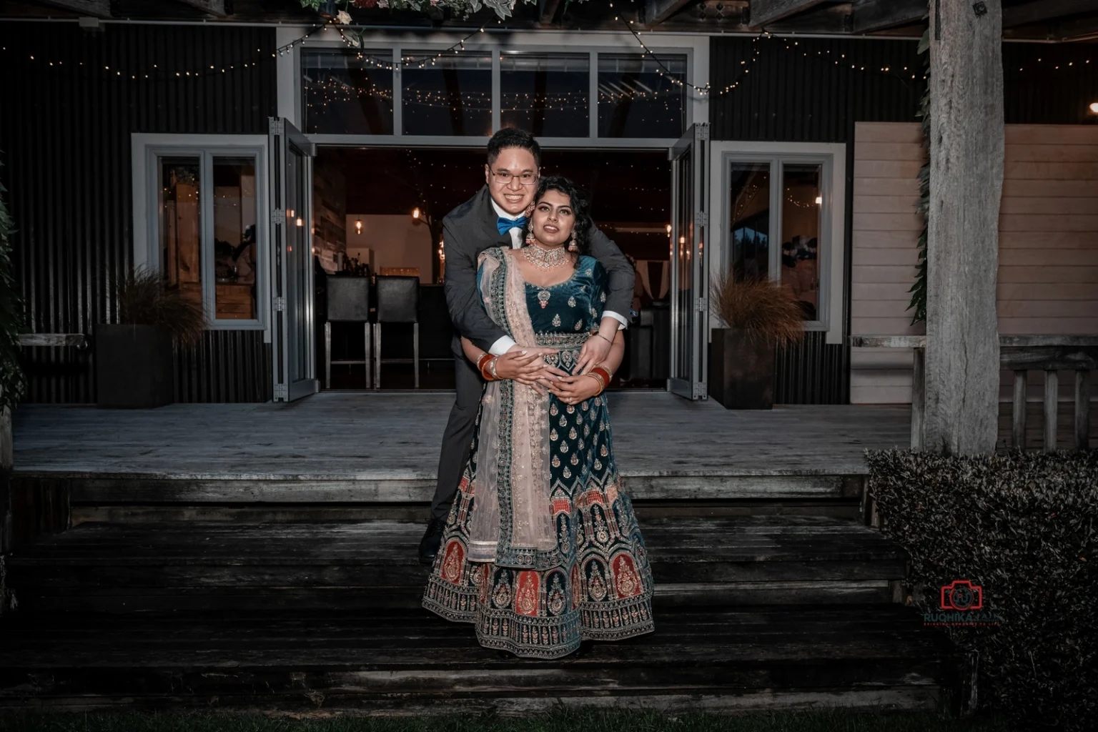 Groom hugs bride from behind as she wears traditional green and gold attire, standing on steps in front of a decorated venue