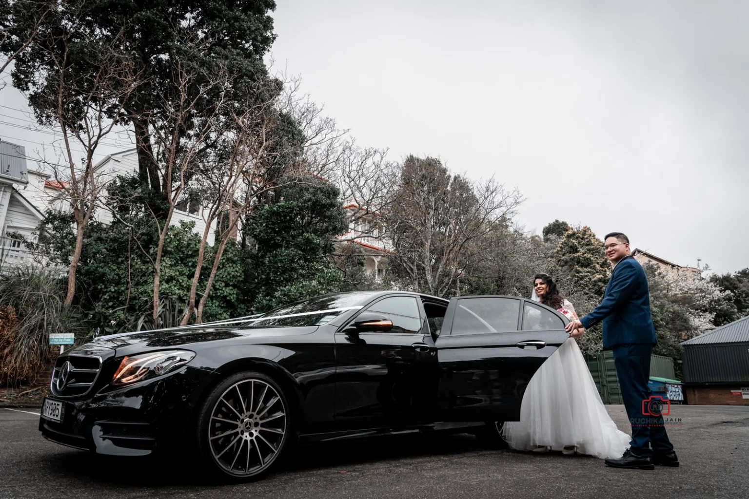 Groom holds open the door of a black Mercedes for the bride, who is seated in the car, with trees and buildings in the background