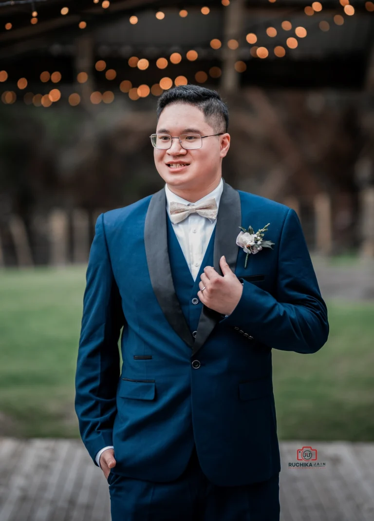 A groom in a stylish navy blue suit and bowtie, standing confidently with a radiant smile at Ohariu Farm wedding venue, with ambient bokeh lights in the background.