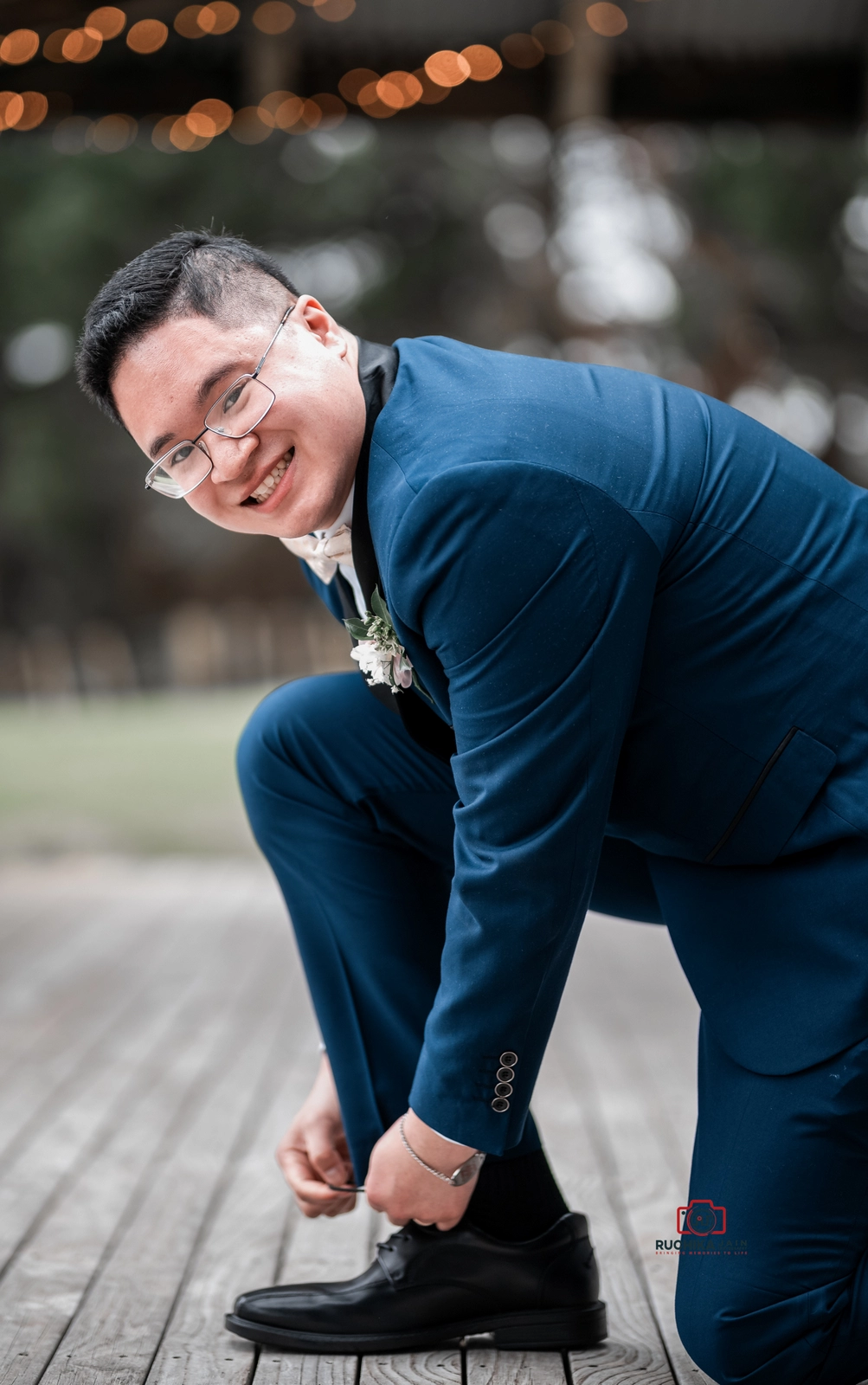 Smiling groom in a blue suit ties his shoe, leaning forward outdoors on a wooden deck with warm string lights in the background