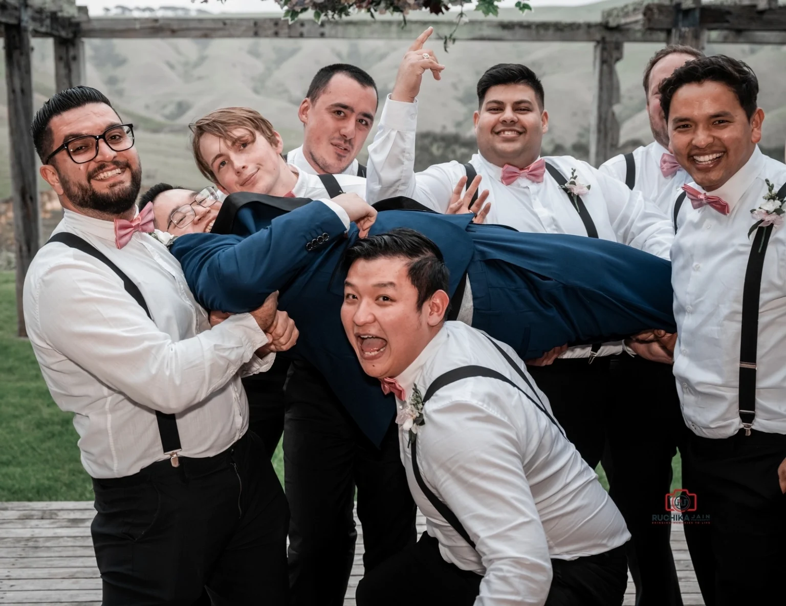 Groomsmen dressed in white shirts and pink bow ties lift the groom in a blue suit, laughing and striking playful poses