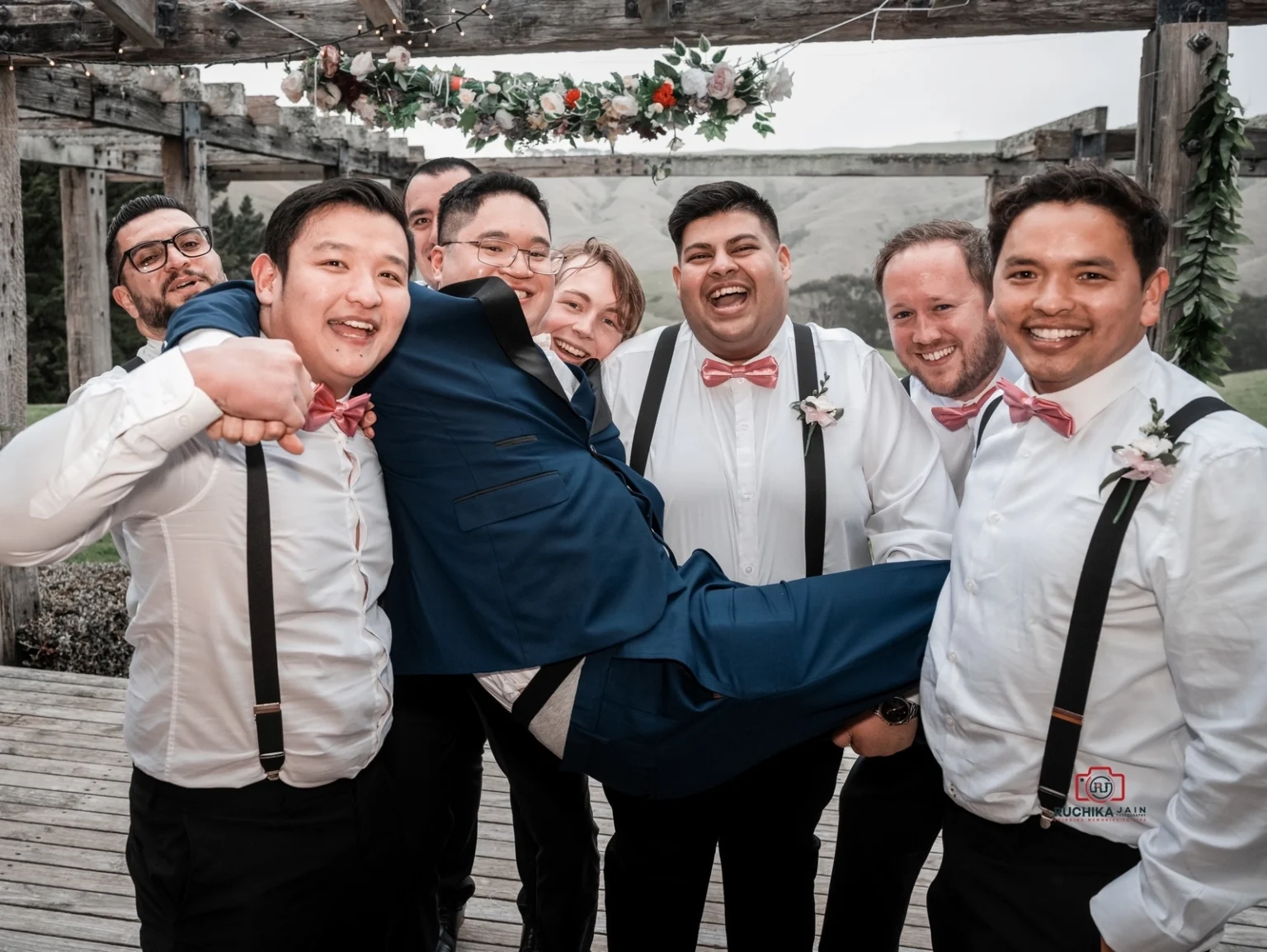 Groomsmen in white shirts and pink bow ties lift the groom in a blue suit, laughing together in a playful wedding photo