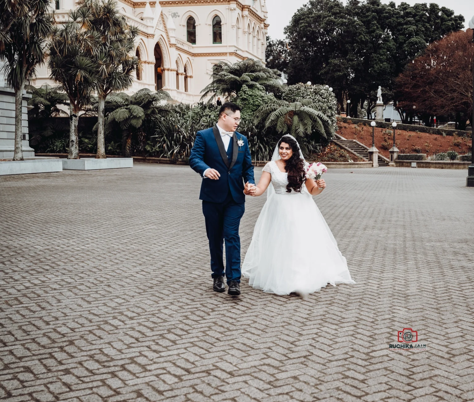 Bride and groom walking hand in hand on a paved pathway outside a historic building, with the bride holding a bouquet and smiling