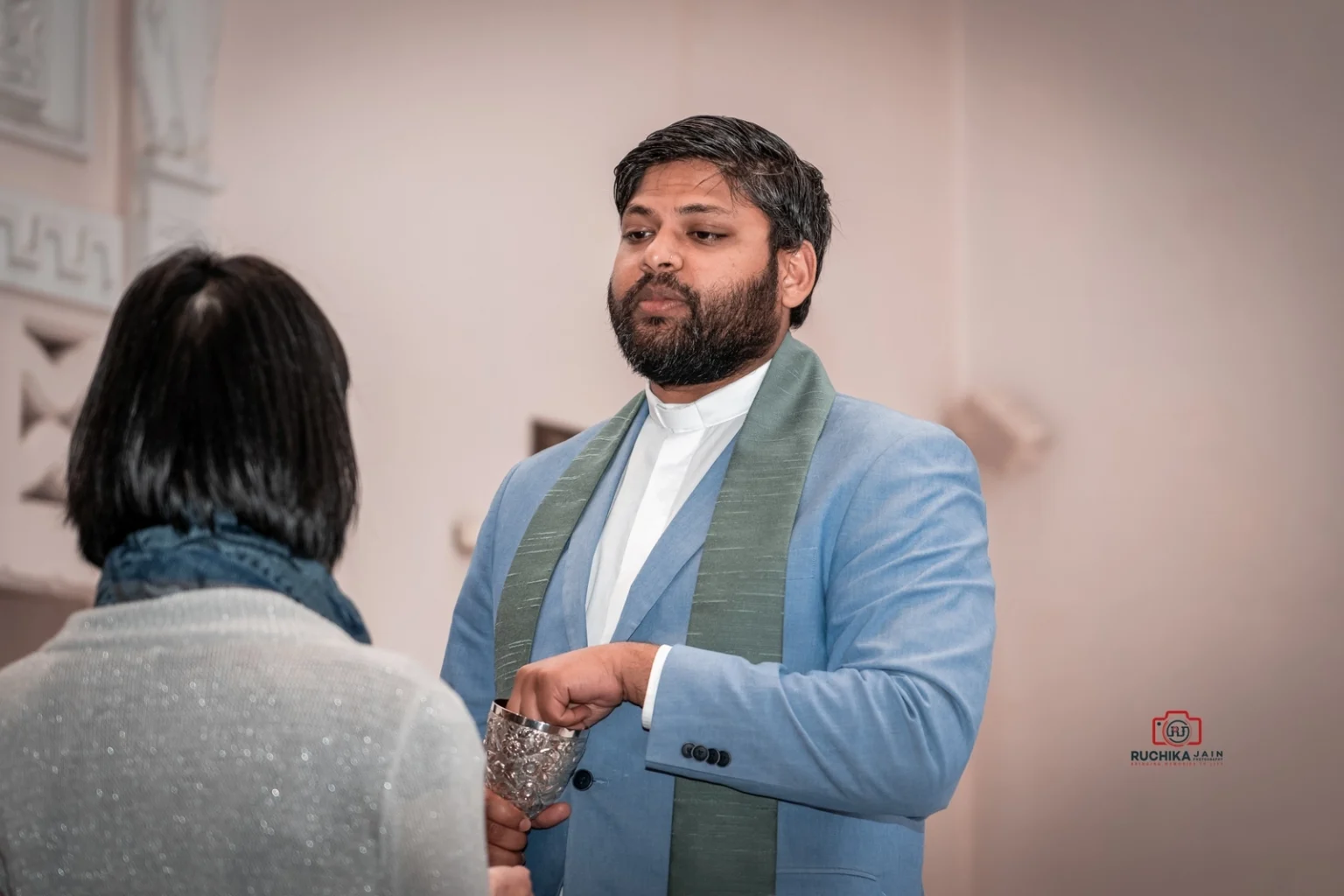 Priest in blue jacket and green stole offering communion to a wedding guest