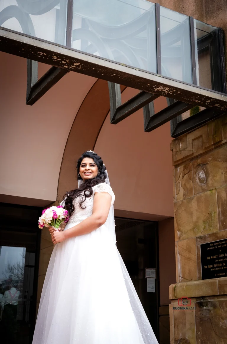 A radiant bride in a white wedding gown holding a pink bouquet, smiling gracefully in front of the Wellington Cathedral of St. Paul