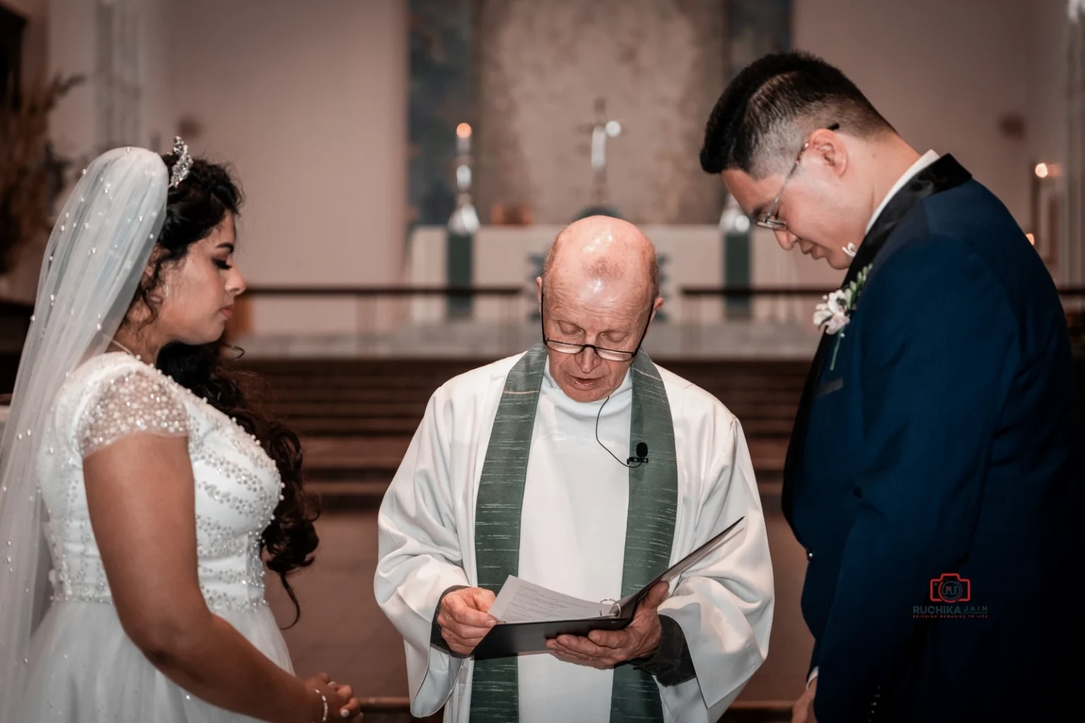 Bride and groom bow their heads during a wedding ceremony led by an officiant holding a book in a church