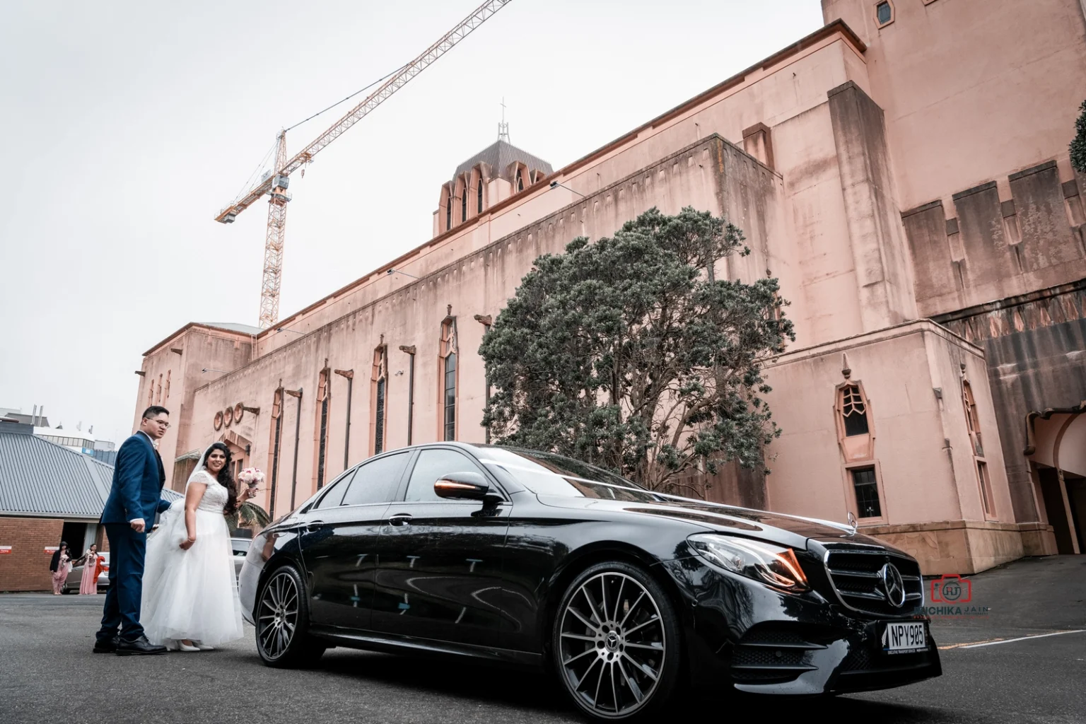 Bride and groom stand beside a black Mercedes sedan in front of a historic building with tall windows and trees, with a crane in the background