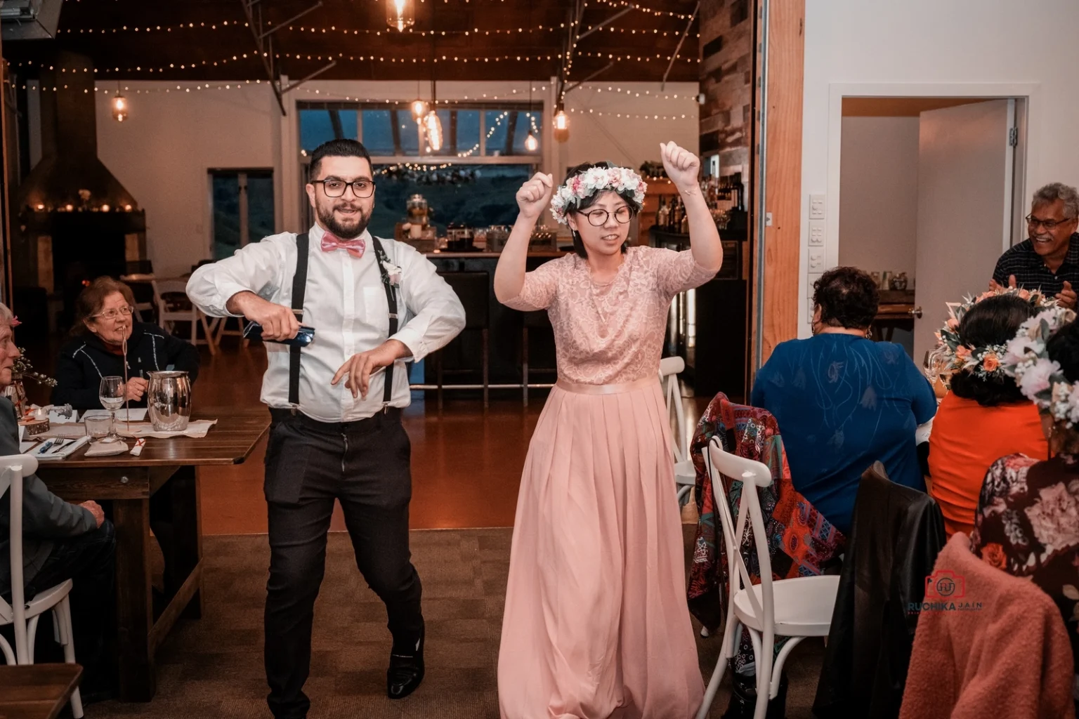 Wedding guests dance joyfully in a warmly lit reception hall decorated with string lights