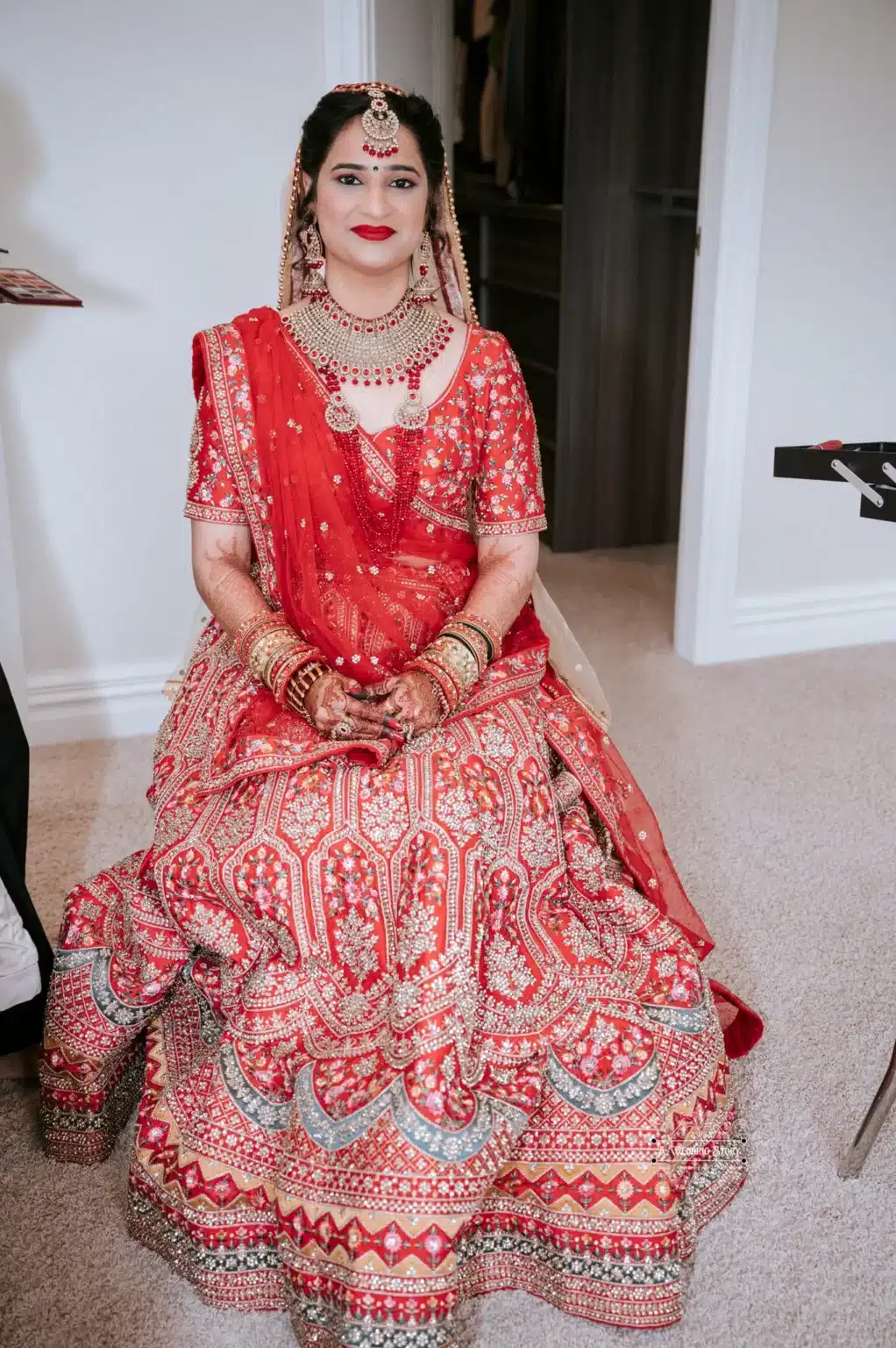 Bride seated in beautiful traditional red attire with intricate embroidery, smiling with hands adorned in mehndi and jewelry.