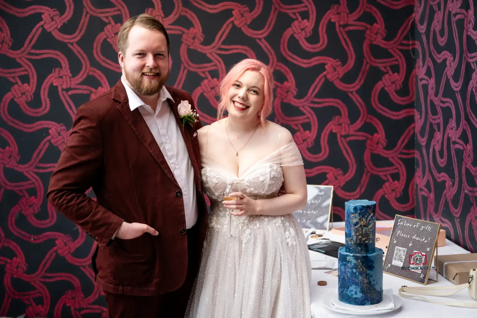 Bride and groom smiling next to their unique blue wedding cake with 'B&G Never Gonna Give You Up' topper