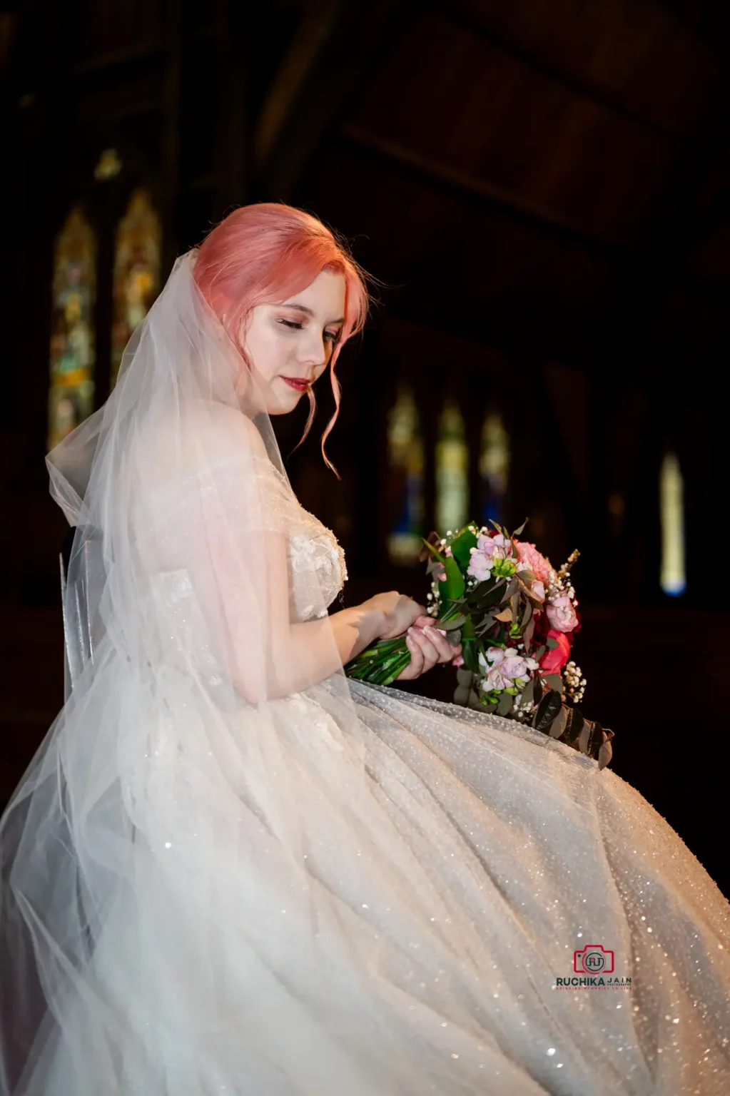 Bride with pink hair holding a bouquet, gazing down, illuminated by soft church lighting.