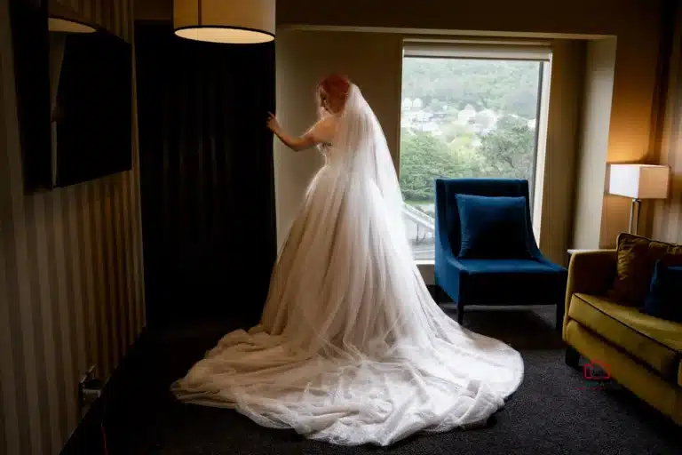 Bride standing in a softly lit room, looking out a window with a long, flowing wedding dress train