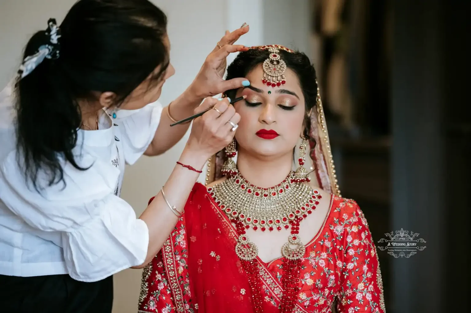 The bride in a red traditional outfit, with intricate jewelry, has her makeup applied before the wedding ceremony in Wellington.