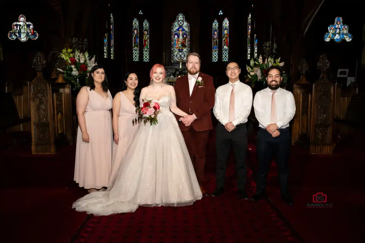 Bride, groom, and their bridal party posing inside a church with stained glass windows.