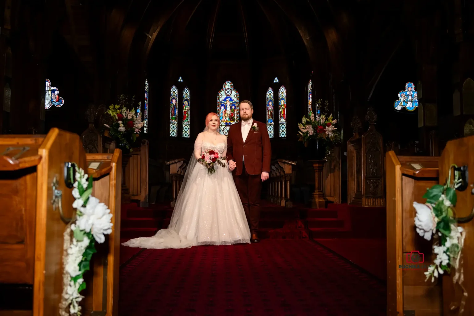 Bride and groom standing hand-in-hand in the church aisle with stained glass windows in the background.