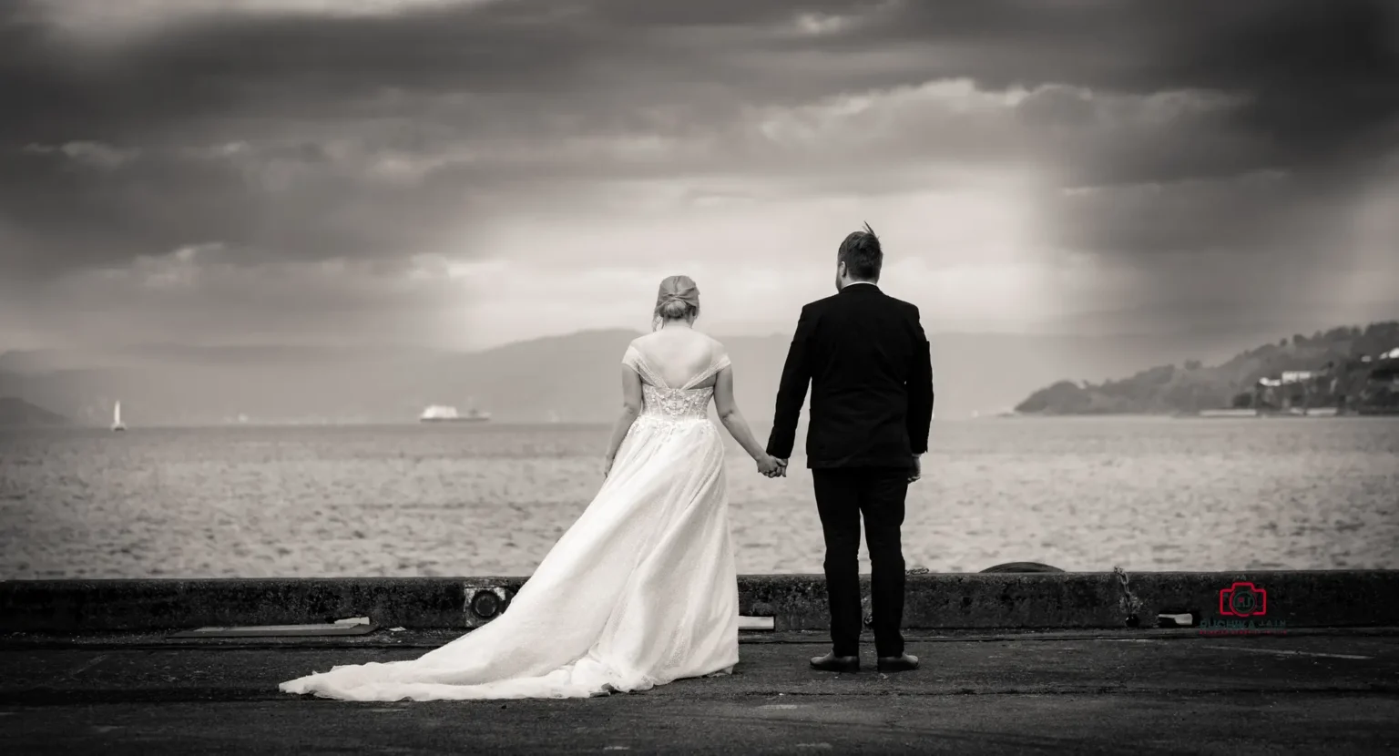 Black and white image of a bride and groom holding hands by the waterfront, facing the ocean with a dramatic cloudy sky overhead.