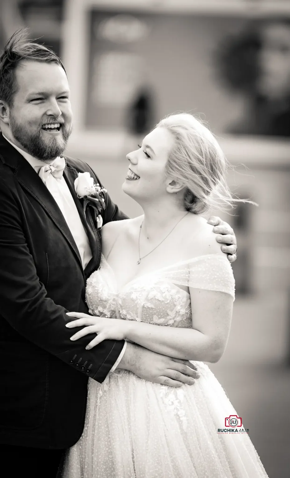 Black and white photo of a bride and groom laughing, with the bride holding the groom and looking up at him