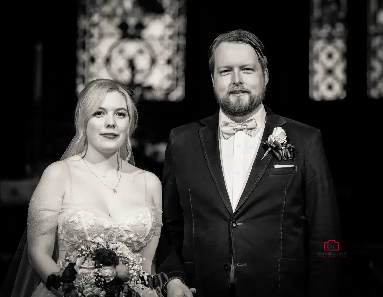 Black and white portrait of the bride and groom standing together at the altar, looking directly at the camera with stained glass in the background.