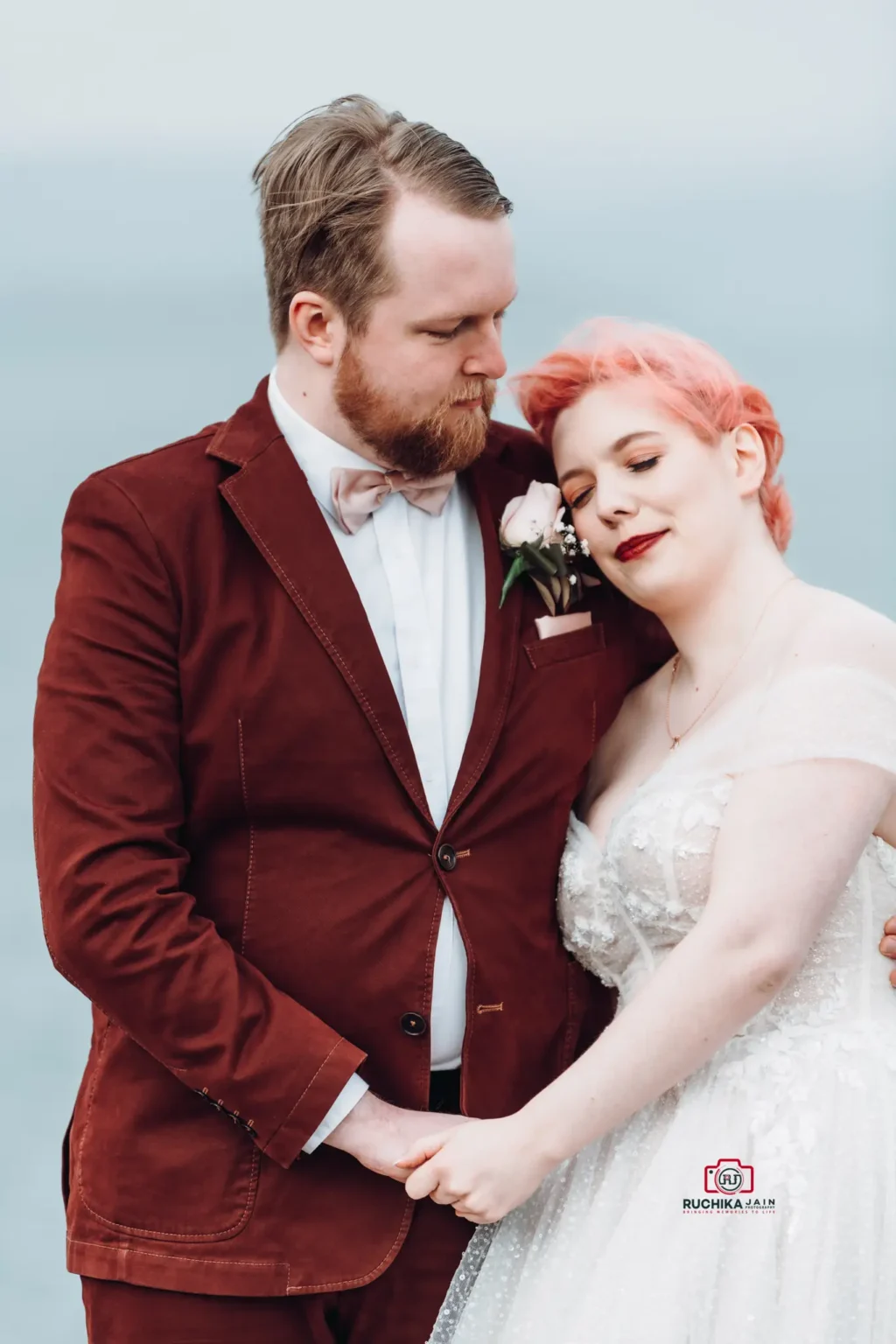 Groom in maroon suit embraces bride with pink hair, holding hands by the seaside with a soft, overcast sky in the background.