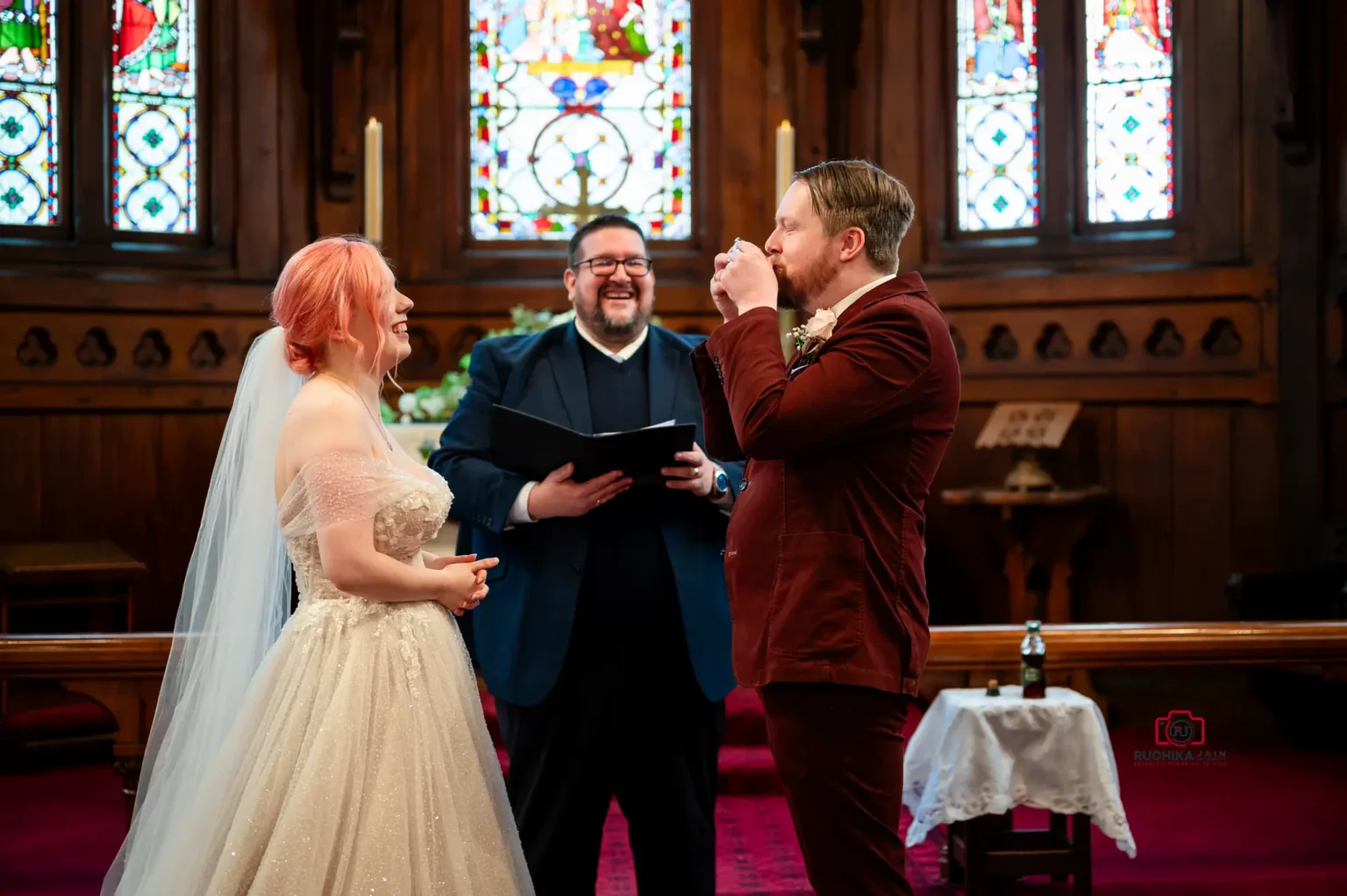 Bride and groom sharing communion during wedding ceremony at a church altar, with officiant smiling and stained glass windows in the background.