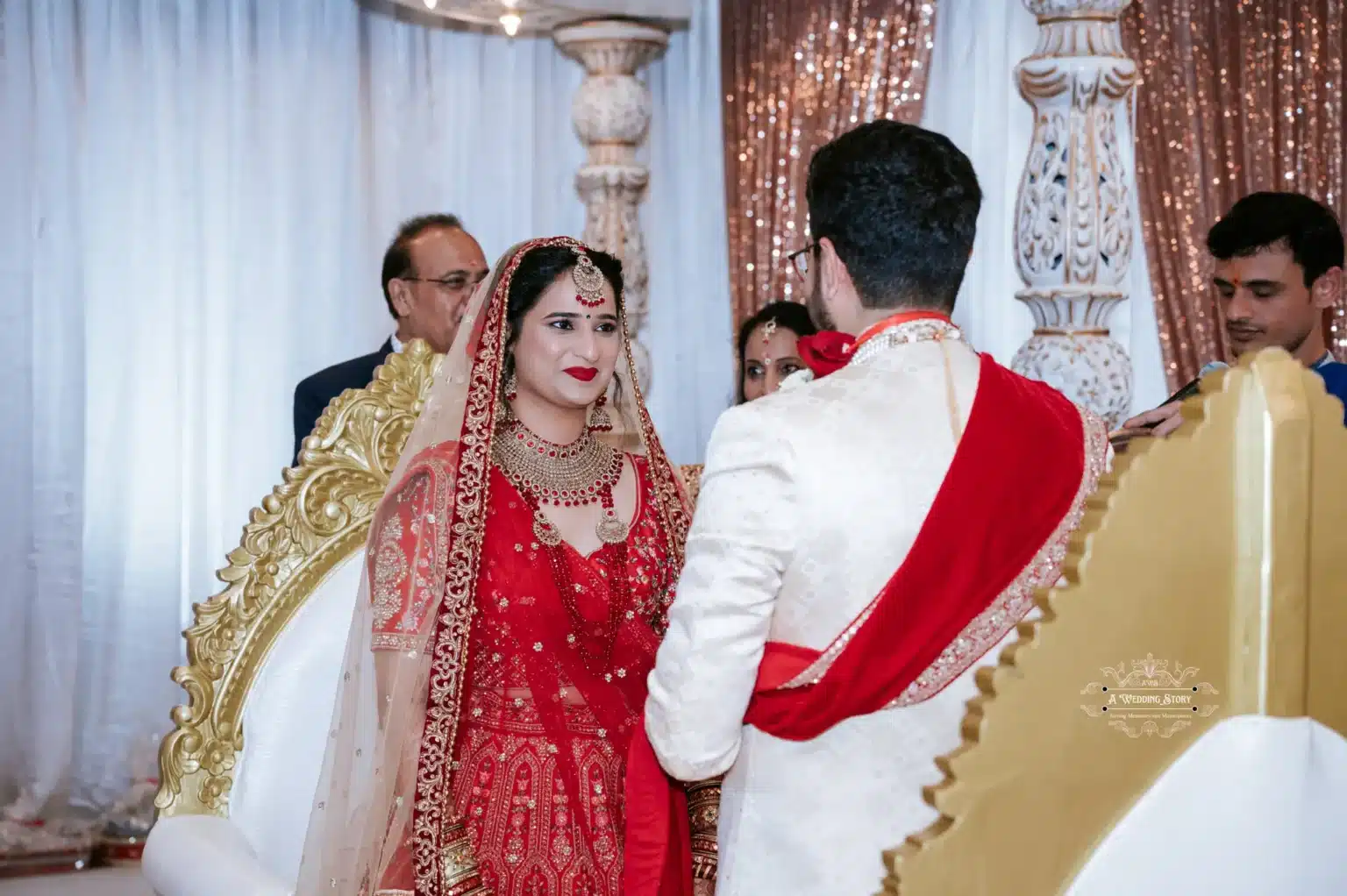 Bride in red attire and groom in white and red outfit exchange smiles during their wedding ceremony.