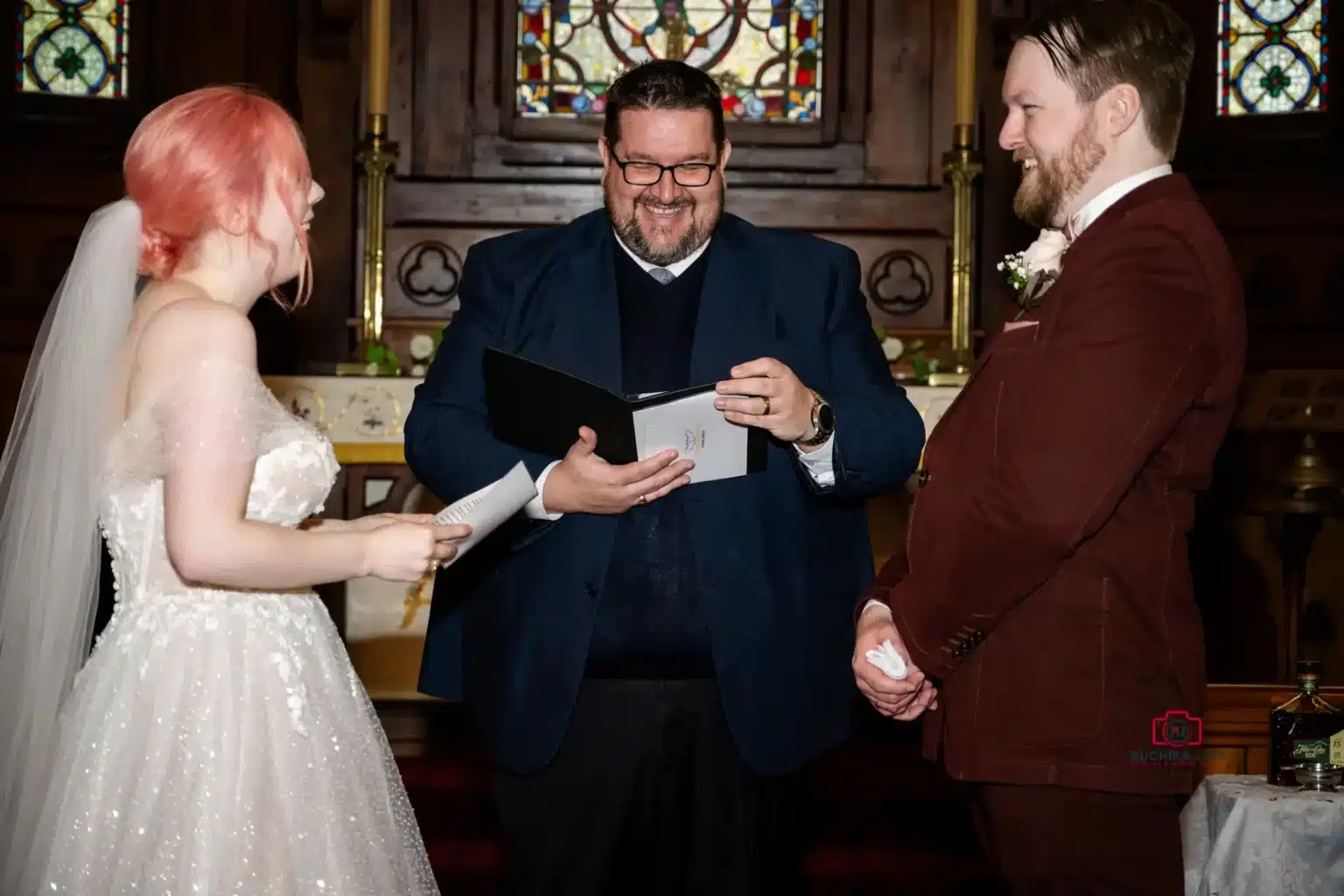 Bride and groom exchange vows while the officiant smiles warmly in a church ceremony.