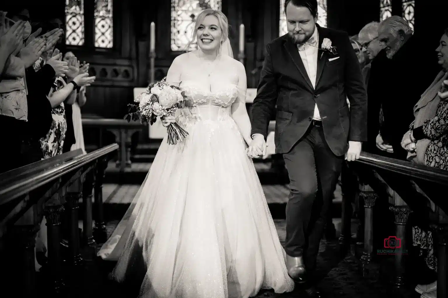 Black and white photo of the bride and groom walking down the aisle after their wedding ceremony, smiling and holding hands.