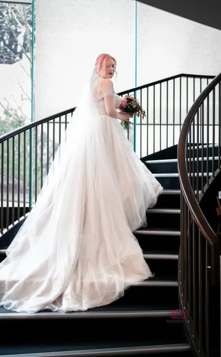 Bride with pink hair and flowing white gown holding a bouquet as she ascends a curved staircase.