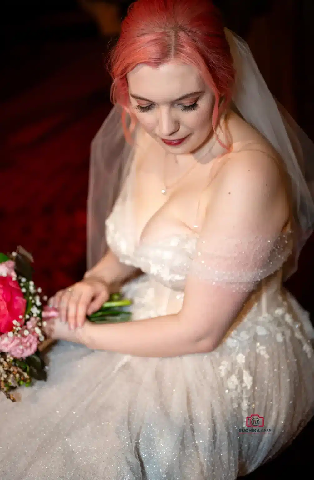 Bride with pink hair, wearing a sparkling gown, looking down at her bouquet.