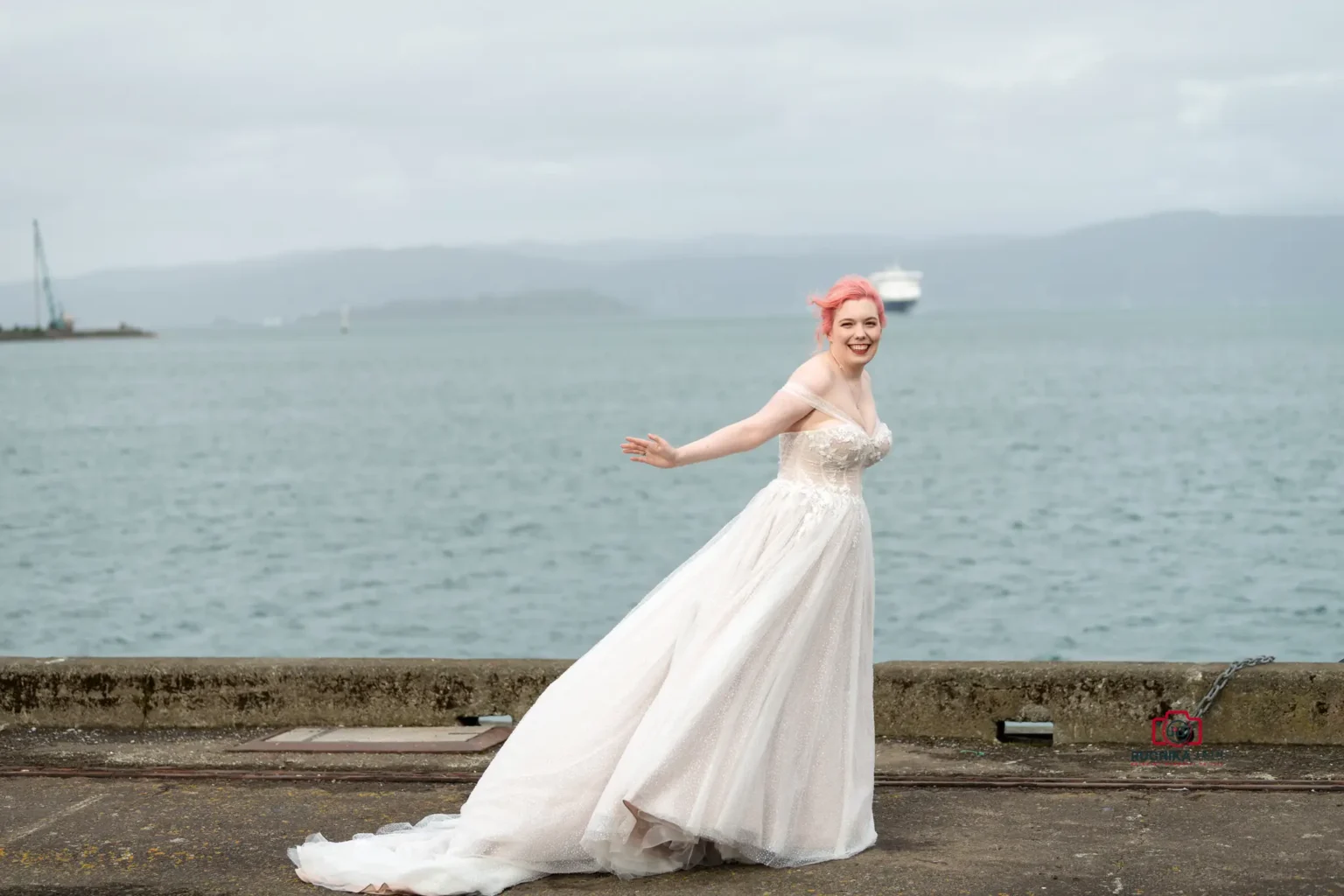 Bride with pink hair in a lace wedding gown, standing by the waterfront with her arms outstretched, smiling at the camera