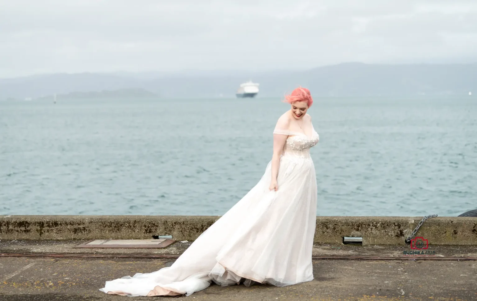 Bride with pink hair in an off-shoulder lace gown, standing by the waterfront with a soft smile, distant mountains, and a boat in the background.