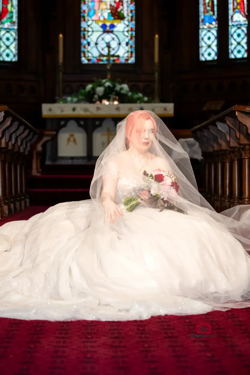 Bride sitting under a flowing veil, holding a bouquet, in a church with stained glass windows.