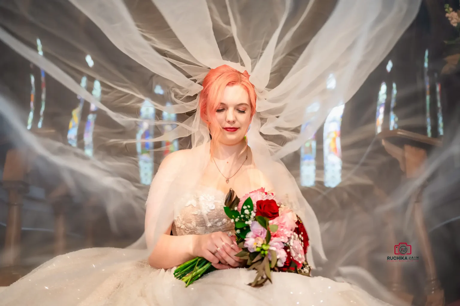 Bride with flowing veil holding a colorful bouquet in front of stained glass windows.