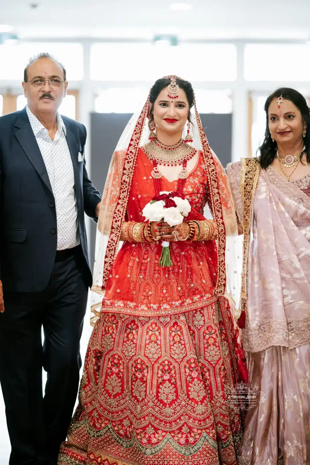 `Bride in traditional red attire holding a bouquet, accompanied by her parents as they walk down the aisle.