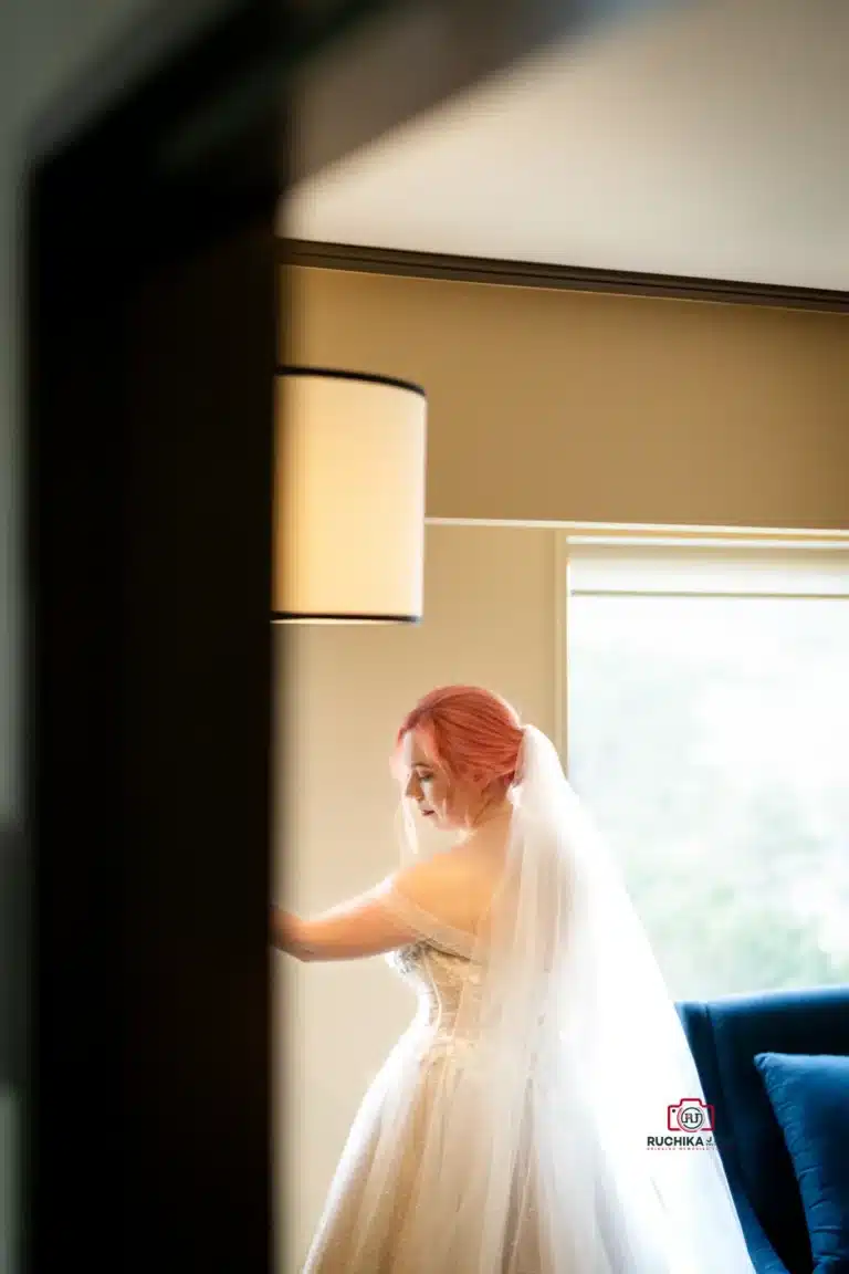 Bride in a delicate moment, softly lit by natural window light as she stands near a lampshade