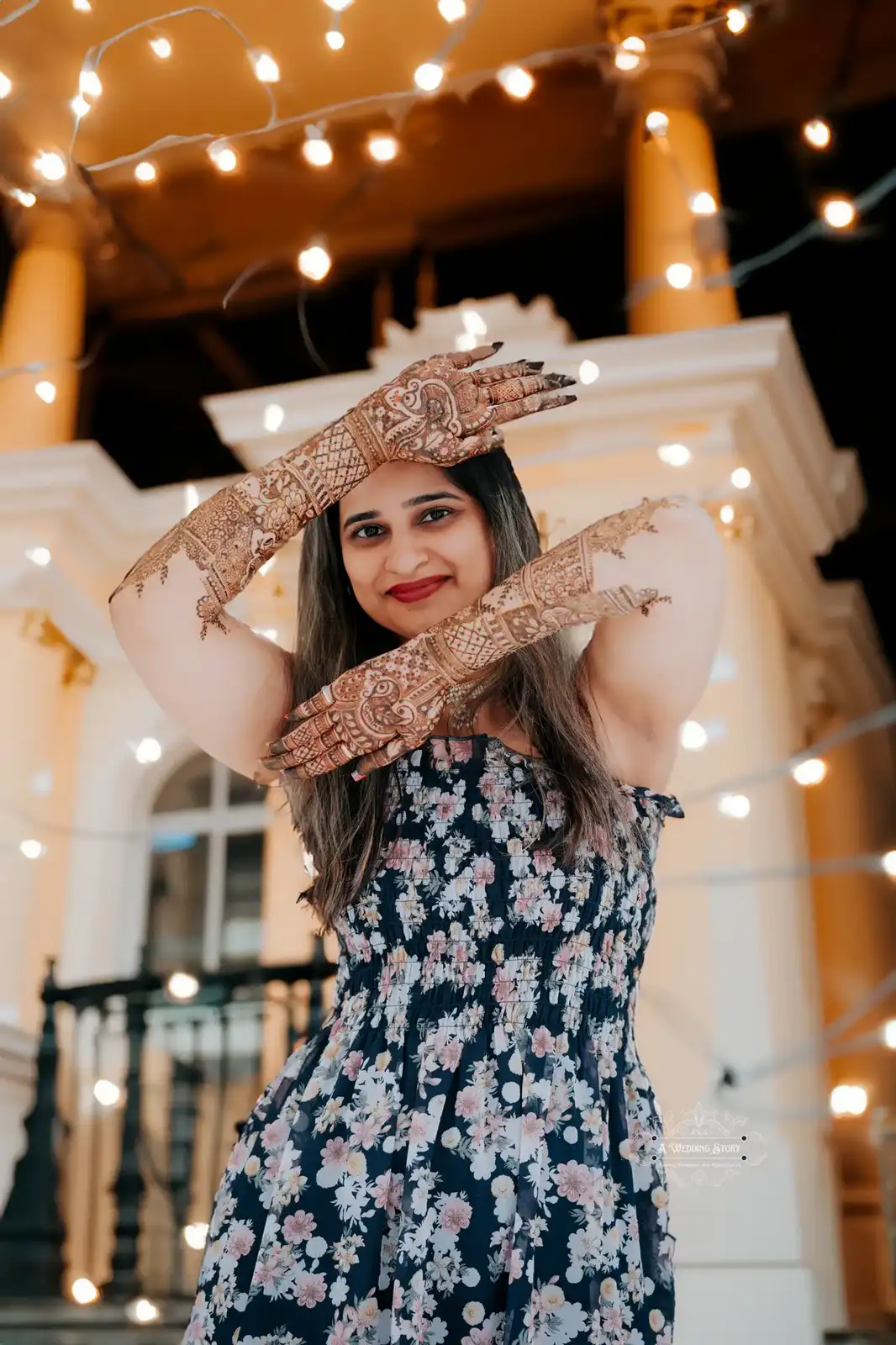 Smiling bride showcasing her bridal mehndi designs under decorative lights, wearing a floral dress, captured during a pre-wedding celebration in Wellington by A Wedding Story