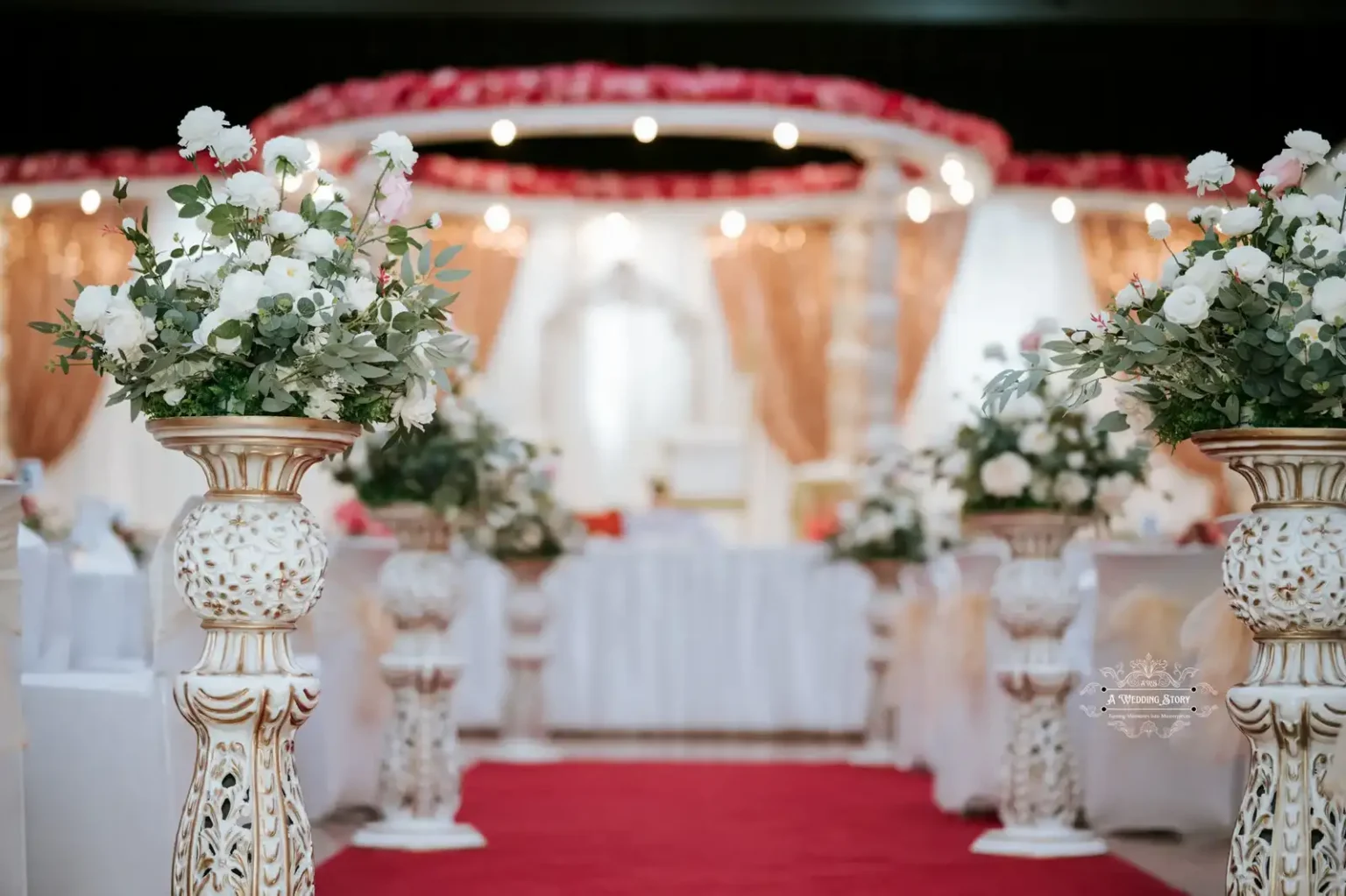 Close-up of an aisle decorated with ornate floral arrangements leading to a wedding stage with white and gold drapery.